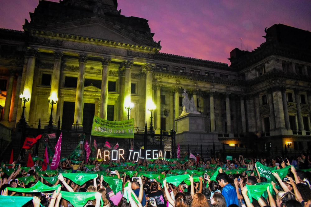 A crowd of people holding up green bandanas in front of a building with columns.