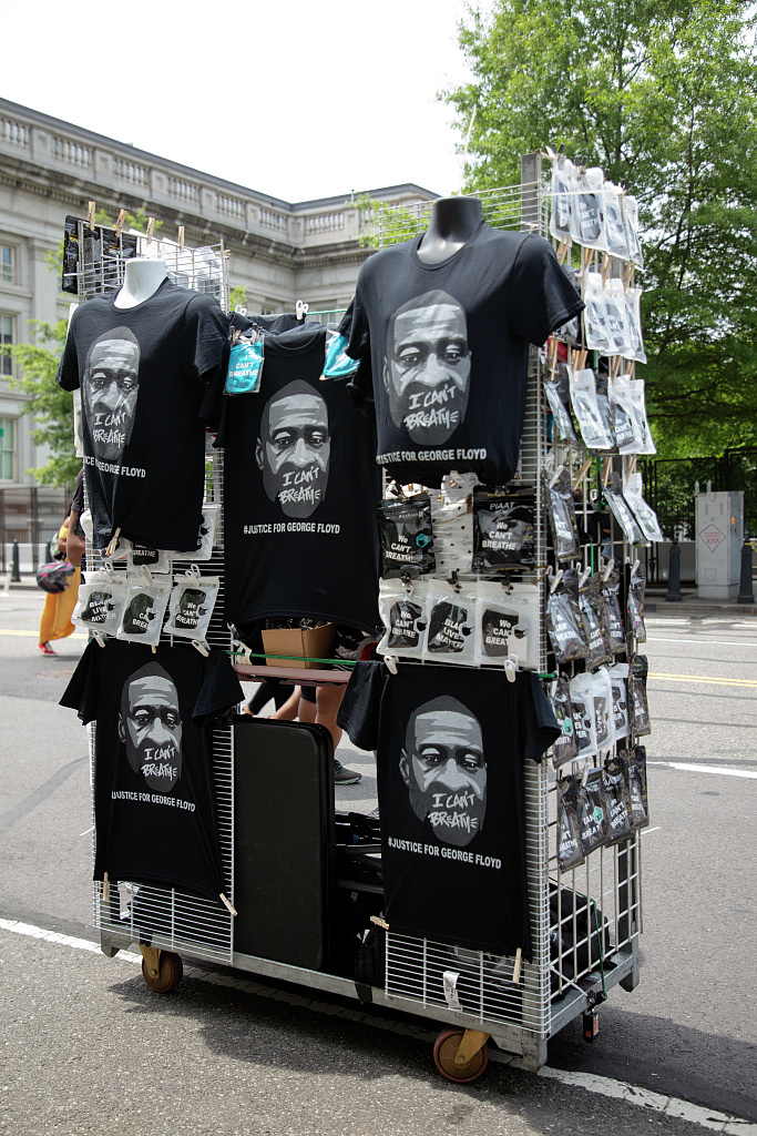 A photograph of a rack of black t-shirts printed with George Floyd’s face, “I CAN’T BREATHE,” and “#JUSTICE FOR GEORGE FLOYD” on the sidewalk at a Washington, D.C. protest.
