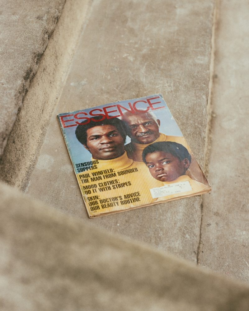 An image of three men in yellow turtlenecks on the cover of a magazine which rests on concrete steps. The magazine has ESSENCE written across the top in red, all-caps, sans serif type. Along the left side of the magazine in black, all-caps, sans serif, condensed type reads: “SENSUOUS SUPPERS, PAUL WINFIELD: THE MAN FROM SOUNDER, MOOD CLOTHES: DO IT WITH STRIPES, SKIN: OUR DOCTOR’S ADVICE OUR BEAUTY ROUTINE.”