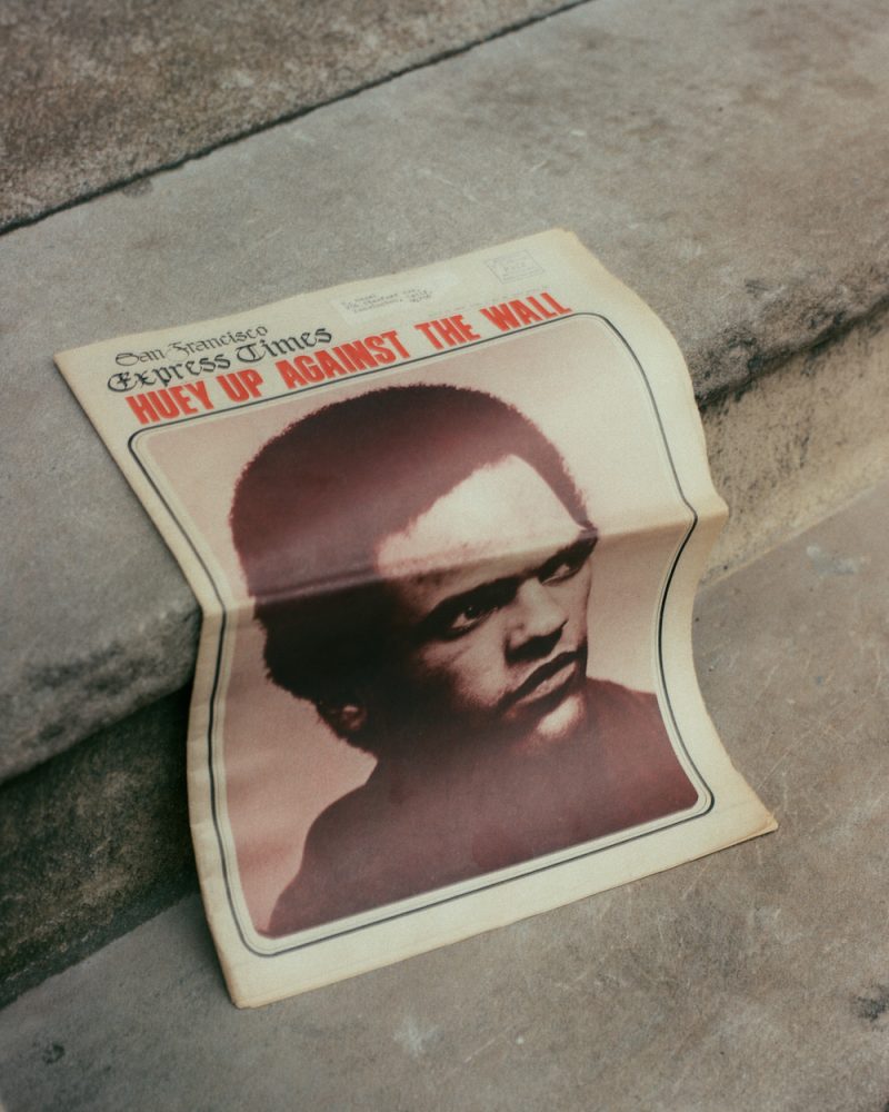 A black-and-white portrait of a man on the cover of a newspaper which rests on concrete steps. The newspaper headline reads, “HUEY UP AGAINST THE WALL” in all-caps, red, sans serif, condensed type.