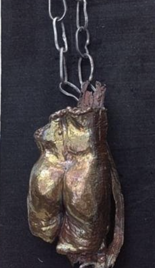 A picture containing a copper form of the lower half of a body shown from the rear, attached to a silver chain, resting on a piece of black cloth against light-colored wood.