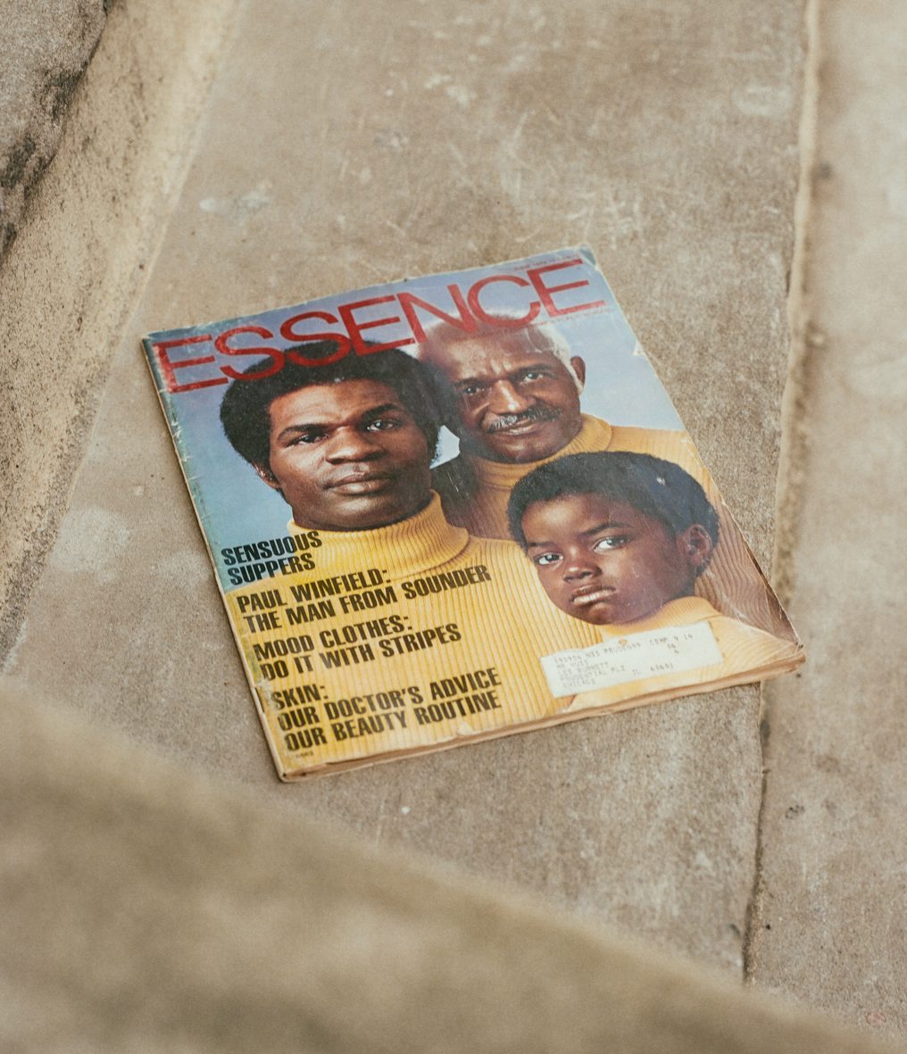 An image of three men in yellow turtlenecks on the cover of a magazine which rests on concrete steps. The magazine has ESSENCE written across the top in red, all-caps, sans serif type. Along the left side of the magazine in black, all-caps, sans serif, condensed type reads: “SENSUOUS SUPPERS, PAUL WINFIELD: THE MAN FROM SOUNDER, MOOD CLOTHES: DO IT WITH STRIPES, SKIN: OUR DOCTOR’S ADVICE OUR BEAUTY ROUTINE.”
